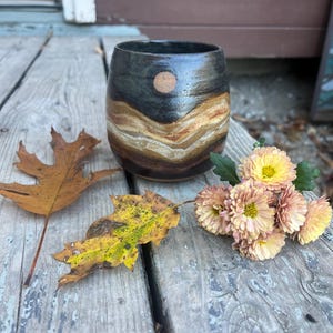 Op de afbeelding: Een handgemaakte keramische beker met een gelaagd ontwerp in tinten zwart, bruin en bruin. Een klein, rond, bruin accent bevindt zich aan de voorkant. De beker wordt gepresenteerd met herfstbladeren en bloemen op een houten oppervlak.