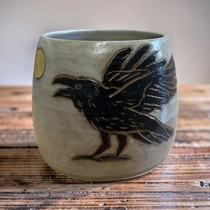 May include: A handmade ceramic mug with a raven design. The mug is a light gray color with a black raven carved into the side. A yellow circle, representing the moon, is also on the side. The mug is on a wooden surface.