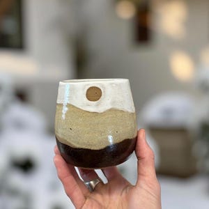 May include: A hand-held, stemless ceramic wine glass with a layered design. The top is white, followed by a tan band, and a dark brown base. A small, round, wooden accent is on the white section.
