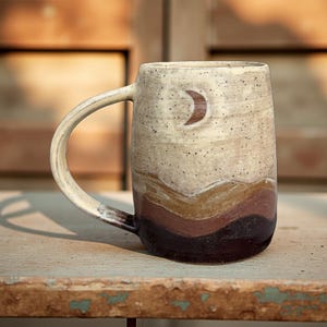Puede incluir: Taza de cerámica hecha a mano con diseño de media luna. La taza presenta una parte superior beige moteada y un diseño de paisaje en capas en tonos marrones y burdeos. El asa es curva y está unida al lateral de la taza.