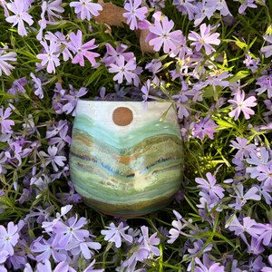 May include: A handmade ceramic tumbler with a landscape design in shades of green, blue, and brown, topped with a brown circle. The tumbler is surrounded by small purple flowers and green foliage.