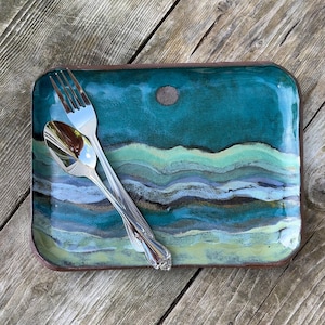 May include: A rectangular ceramic tray with a glossy glaze featuring horizontal wavy bands of teal, light green, blue, and yellow, with a small circular brown accent. A polished silver-toned metal fork and spoon rest diagonally on the tray surface.