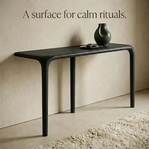 Pode incluir: Uma mesa consola preta com bordas arredondadas e três pernas, apresentando um vaso escuro e um pequeno prato em cima. O texto "A surface for calm rituals." é exibido acima da mesa, que está sobre um piso de cor clara.