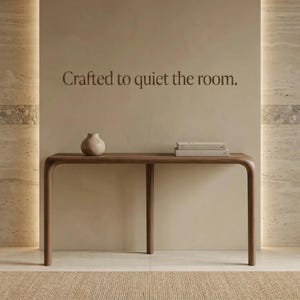 Peut inclure: Une console en bois fonc&eacute; aux bords arrondis, surmont&eacute;e d'un petit vase en c&eacute;ramique beige et de deux livres empil&eacute;s. Le mur au-dessus de la table affiche le texte brun fonc&eacute;, Crafted to quiet the room, dans une police &agrave; empattements.