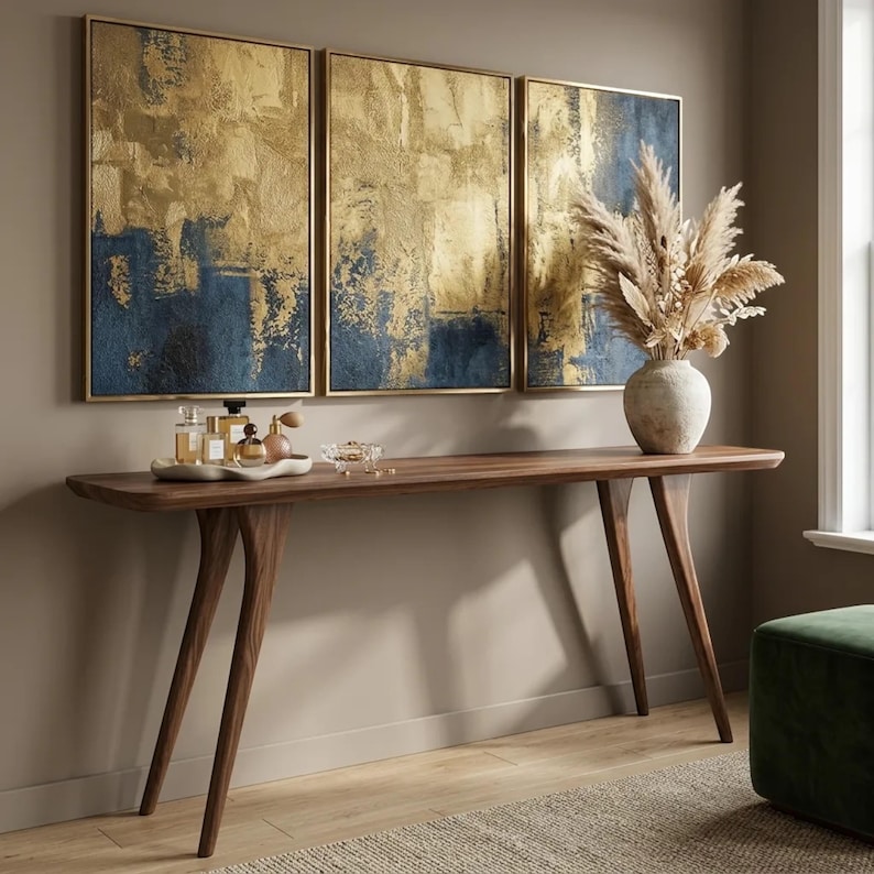 May include: A long wooden console table with four tapered legs holds a white tray with glass perfume bottles and a textured vase filled with dried pampas grass. Above the table hang three rectangular canvases featuring abstract gold and navy blue paint.