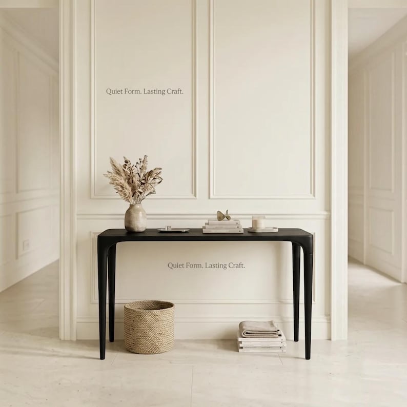 Puede incluir: Una mesa consola negra con cuatro patas c&oacute;nicas, con un jarr&oacute;n claro con plantas secas, una pila de libros y una vela encima. Debajo hay una cesta tejida y una pila de textiles doblados. Aparece el texto "Quiet Form. Lasting Craft.".