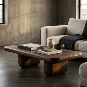 May include: A dark wood coffee table with a rectangular top and rounded edges, supported by two curved legs. Two books, a small white vase, and a black bowl are on the table. A beige sofa and concrete wall are in the background.