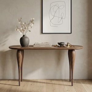 Può includere: Un tavolo consolle in legno con piano ovale e quattro gambe curve. Sul tavolo si trovano un vaso grigio scuro con steli essiccati, due libri impilati intitolati Japandi Living e Atelier, e un vassoio dorato con una ciotola e un campanello in ottone.