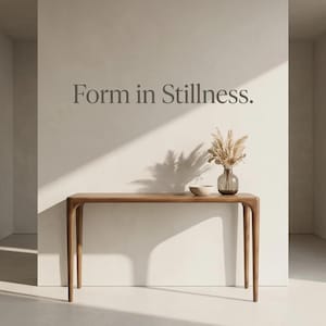 May include: A wooden console table with rounded legs stands against a wall featuring the text Form in Stillness. On the table sits a small beige bowl and a glass vase containing dried pampas grass, illuminated by a diagonal beam of sunlight.