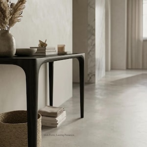 Puede incluir: Una mesa consola de madera oscura con cuatro patas delgadas y una superficie rectangular lisa. Sobre la mesa hay un jarr&oacute;n de cer&aacute;mica con hierbas secas, un cuenco peque&ntilde;o, una pila de libros y una vela. El texto Slim Form, Lasting Presence es visible.