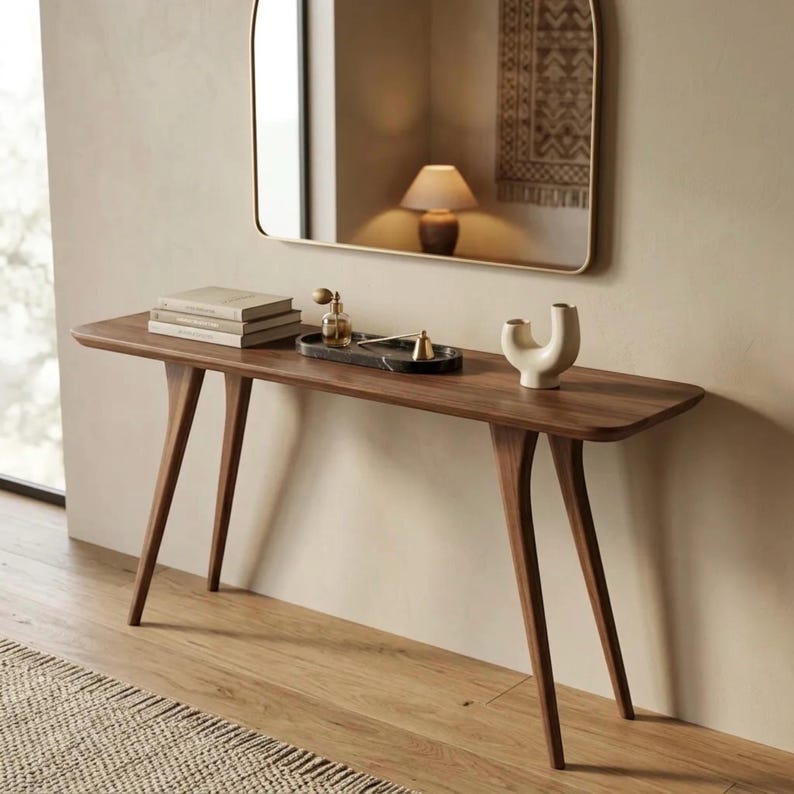 May include: A long, narrow wooden console table with four tapered legs. On the surface sits a stack of three books, a black marble tray holding a glass perfume bottle and a brass object, and a cream-coloured ceramic sculpture with a curved shape.