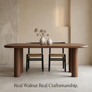 May include: A walnut dining table with a smooth, rounded rectangular top and four thick cylindrical legs. Two black chairs with woven seats are positioned at the table. Two white ceramic vases hold dried flowers. Text reads: Real Walnut Real Craftsmanship.