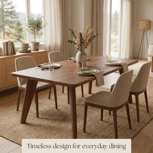 May include: A rectangular wooden dining table with four upholstered chairs in a light beige fabric. The table is set with plates, glasses, and napkins. A ceramic vase with dried plants sits in the centre. Text reads: Timeless design for everyday dining.
