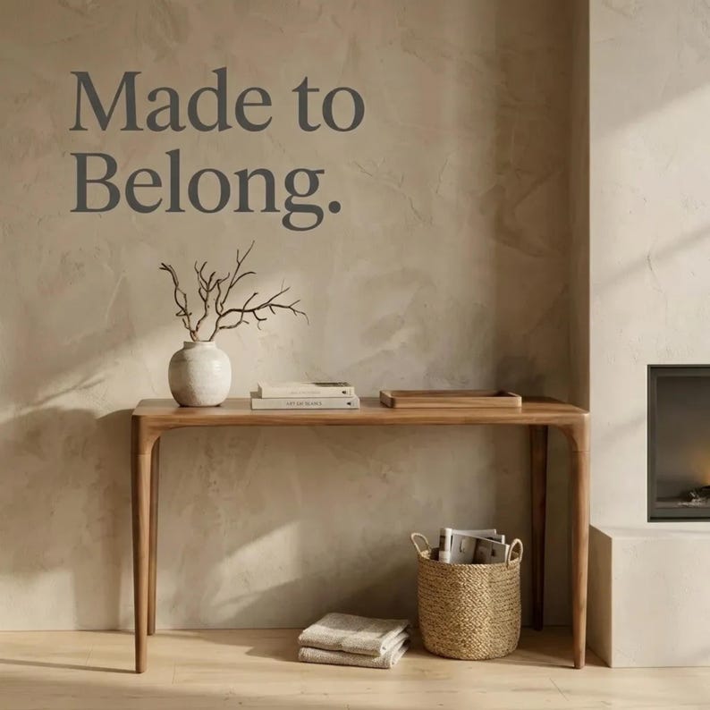 May include: A wooden console table with rounded legs, topped with a white vase containing bare branches, two stacked books, and a wooden tray. The wall features the text Made to Belong. A woven basket with magazines and folded textiles sits underneath.
