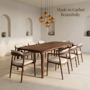 Può includere: Un tavolo da pranzo in legno scuro con bordi arrotondati e otto sedie abbinate con sedute imbottite bianche. Sopra il tavolo pende un gruppo di cinque lampade a sospensione in vetro ambrato. Il testo "Made to Gather Beautifully" è visibile.