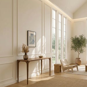 May include: A modern living room with a wooden console table, a framed abstract art piece, and a vase with dried flowers. The room features large windows, a woven rug, a wooden chair, and a potted olive tree, creating a bright and airy atmosphere.
