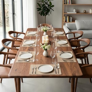 Peut inclure: Une table à manger rectangulaire en bois de noyer dressée pour six personnes avec des assiettes en céramique claire, des couverts en argent, des verres à pied transparents et des sets de table tissés. Deux vases blancs avec des fleurs.