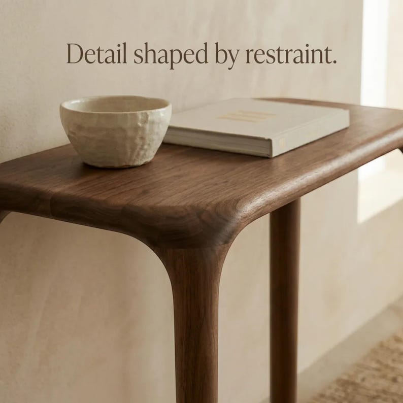 Peut inclure: Une console en bois fonc&eacute; aux bords arrondis, surmont&eacute;e d'un bol en c&eacute;ramique blanc cass&eacute; textur&eacute; et d'un livre ferm&eacute; de couleur claire. Le texte "Detail shaped by restraint." est imprim&eacute; en lettres marron fonc&eacute; au-dessus de la table.