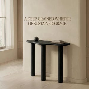 Puede incluir: Una mesa consola negra con tres patas cilíndricas y una superficie de forma orgánica irregular. Sobre la mesa hay un pequeño jarrón oscuro y una pila de dos libros. En la pared superior aparece el texto: A DEEP-GRAINED WHISPER OF SUSTAINED GRACE.