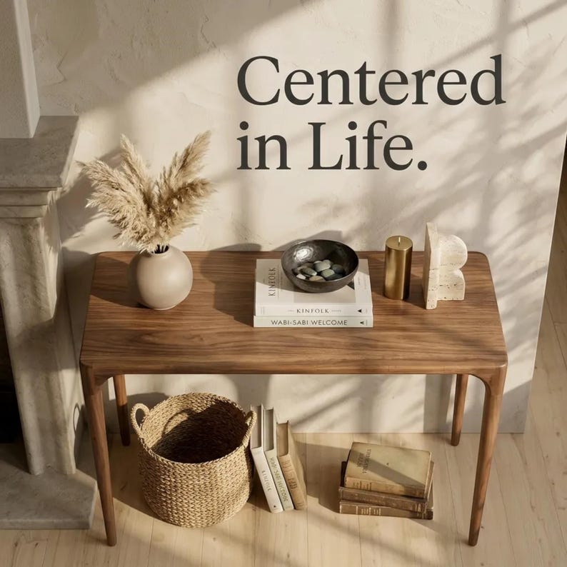 May include: A rectangular wooden console table with rounded edges. On top sits a beige vase with dried grass, a dark bowl with stones, two stacked books titled Kinfolk and Wabi-Sabi Welcome, a gold candle, and a stone sculpture. Text reads Centered in Life.