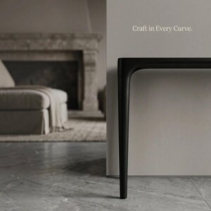 Puede incluir: Una mesa consola negra con una pata y un borde de tablero curvos y suaves. El texto "Craft in Every Curve." es visible en la pared sobre la mesa. El mueble presenta un acabado mate y un perfil de dise&ntilde;o elegante y continuo.