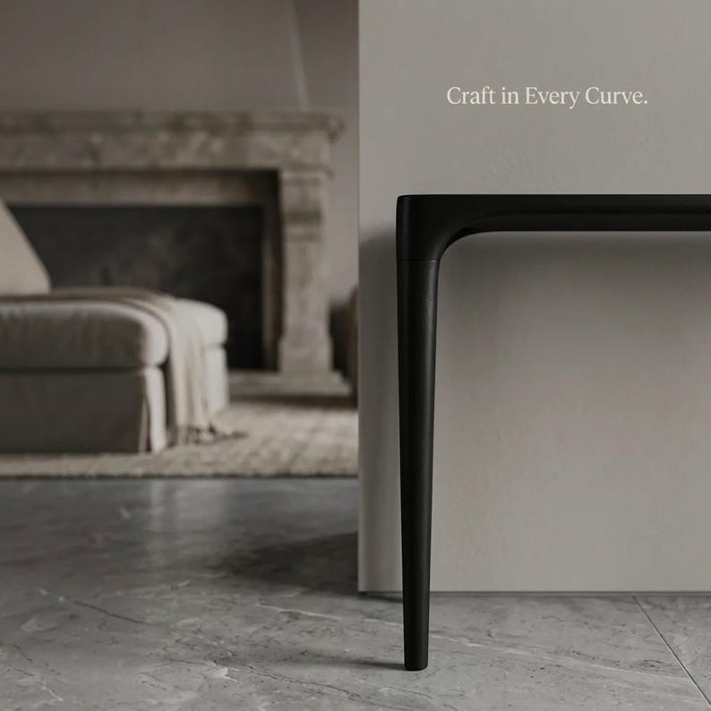 Puede incluir: Una mesa consola negra con una pata y un borde de tablero curvos y suaves. El texto "Craft in Every Curve." es visible en la pared sobre la mesa. El mueble presenta un acabado mate y un perfil de dise&ntilde;o elegante y continuo.