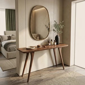 May include: A dark wood console table with four tapered legs stands against a wall. On the table are a small white dish, a wooden brush, three glass perfume bottles, and a black vase with green leaves. Above sits an oval mirror with a gold frame.