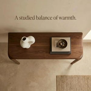 Peut inclure: Une console en bois fonc&eacute; aux bords arrondis, surmont&eacute;e d'un vase en c&eacute;ramique blanche, d'une pile de livres et d'un petit bol sombre contenant des pierres. Le texte "A studied balance of warmth." est inscrit sur le mur au-dessus.
