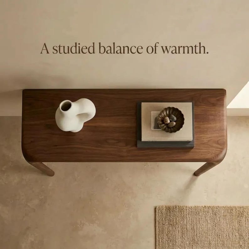 Peut inclure: Une console en bois fonc&eacute; aux bords arrondis, surmont&eacute;e d'un vase en c&eacute;ramique blanche, d'une pile de livres et d'un petit bol sombre contenant des pierres. Le texte "A studied balance of warmth." est inscrit sur le mur au-dessus.