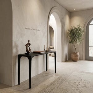 Puede incluir: Una mesa consola negra con cuatro patas delgadas, que presenta una escultura abstracta de bronce, un libro marr&oacute;n y tres velas finas encima. La pared sobre la mesa muestra el texto Crafted to Welcome Well en una fuente oscura y sencilla.