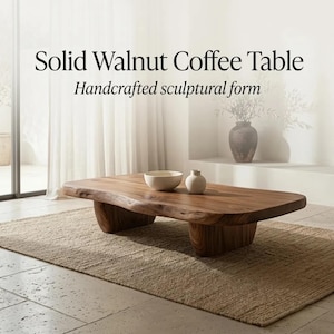 May include: A low-profile solid walnut coffee table with a natural live edge and two thick, rounded wooden supports. The table surface features a rich brown wood grain. Text on the image reads: Solid Walnut Coffee Table Handcrafted sculptural form.