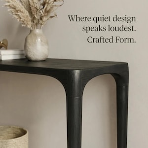 Puede incluir: Una mesa consola de madera negra con patas curvas y una textura de veta de madera visible. Sobre la mesa hay un jarr&oacute;n de cer&aacute;mica de color claro que contiene hierbas secas. El texto "Where quiet design speaks loudest. Crafted Form." es visible.