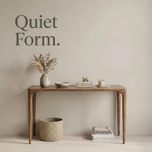 May include: A wooden console table with four tapered legs. On the table are a ceramic vase with dried grasses, a shallow bowl, a stack of books with a small gold object, and a candle. Below sits a woven basket and a stack of folded textiles.