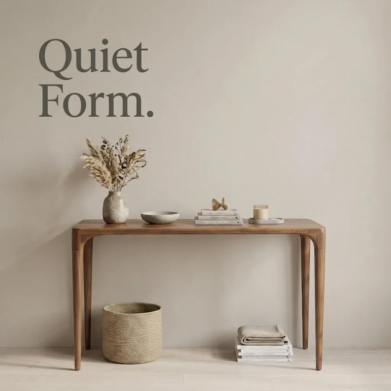 May include: A wooden console table with four tapered legs. On the table are a ceramic vase with dried grasses, a shallow bowl, a stack of books with a small gold object, and a candle. Below sits a woven basket and a stack of folded textiles.