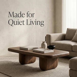 May include: A low, rectangular wooden coffee table with rounded edges and two thick, curved pedestal bases. On the table sit two books, a small beige ceramic vase, and a black bowl. The text "Made for Quiet Living" is visible on the wall.