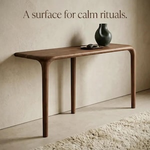 Peut inclure: Une console en bois brun aux bords arrondis et quatre pieds, surmont&eacute;e d'un vase en c&eacute;ramique gris fonc&eacute; et d'un petit plateau sombre. Le texte "A surface for calm rituals." est imprim&eacute; au-dessus de la table en police serif brun fonc&eacute;.