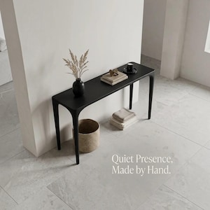 Puede incluir: Una mesa consola negra con cuatro patas delgadas, que presenta un jarr&oacute;n negro con hierbas secas, una pila de libros y una peque&ntilde;a vela negra. El texto "Quiet Presence, Made by Hand." es visible en la esquina inferior derecha.