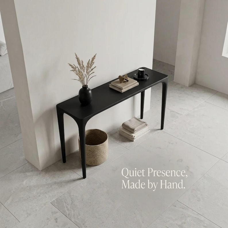 Puede incluir: Una mesa consola negra con cuatro patas delgadas, que presenta un jarr&oacute;n negro con hierbas secas, una pila de libros y una peque&ntilde;a vela negra. El texto "Quiet Presence, Made by Hand." es visible en la esquina inferior derecha.