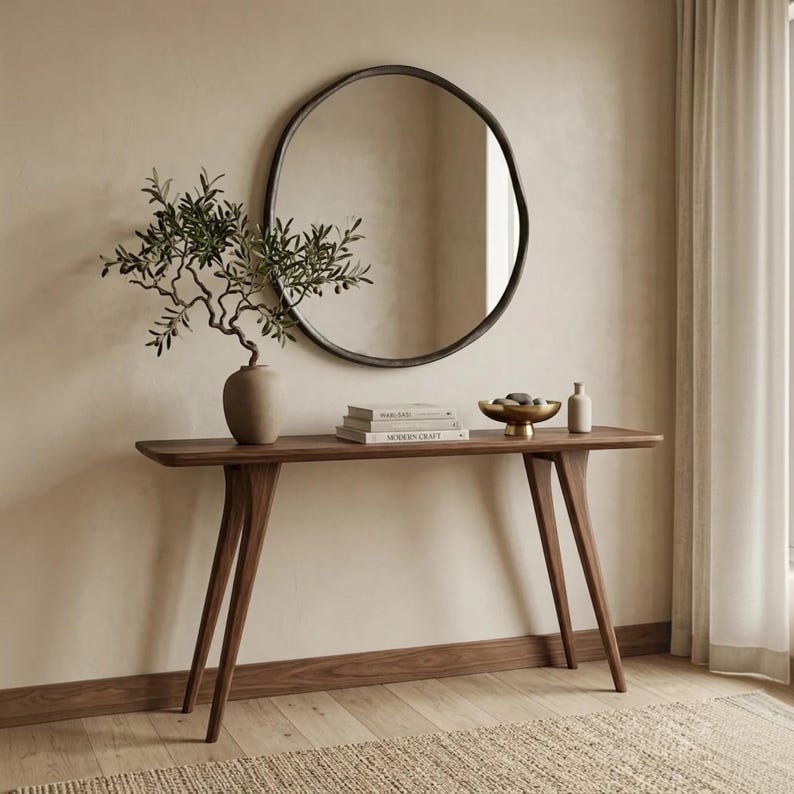 May include: A dark wood console table with four angled legs, topped with a matte beige vase containing an olive branch, a stack of two books, a gold-toned pedestal bowl with stones, and a small bottle, beneath a large round mirror.