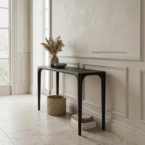 Puede incluir: Una mesa consola negra con cuatro patas delgadas y una superficie rectangular lisa. Un jarr&oacute;n de cer&aacute;mica con flores secas se encuentra sobre la mesa. Una cesta tejida y una pila de libros est&aacute;n colocadas debajo. El texto "Designed for Beautiful Entryways" es visible.