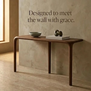 Peut inclure: Une console en bois fonc&eacute; aux bords arrondis et quatre pieds. Sur la table se trouvent un bol gris clair et un livre rectangulaire blanc surmont&eacute; d'une sculpture abstraite sombre. Le mur porte le texte : Designed to meet the wall with grace.