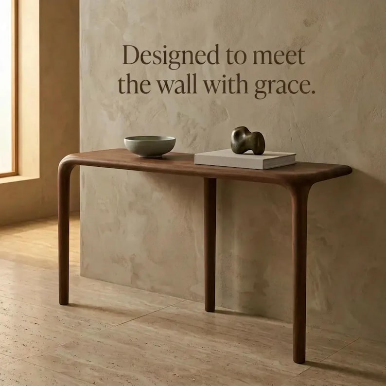 Peut inclure: Une console en bois fonc&eacute; aux bords arrondis et quatre pieds. Sur la table se trouvent un bol gris clair et un livre rectangulaire blanc surmont&eacute; d'une sculpture abstraite sombre. Le mur porte le texte : Designed to meet the wall with grace.