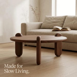 Puede incluir: Una mesa de centro redonda de madera marrón oscuro con una superficie lisa y cuatro patas cilíndricas gruesas que sobresalen ligeramente por encima del tablero. El texto "Made for Slow Living." está impreso en blanco en la esquina inferior izquierda.