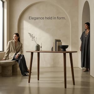 Pode incluir: Uma mesa console de madeira com quatro pernas cônicas está em uma sala com uma parede arqueada apresentando o texto Elegance held in form. Sobre a mesa há um vaso verde claro com um ramo de planta, uma vela fina, três livros e uma tigela preta.