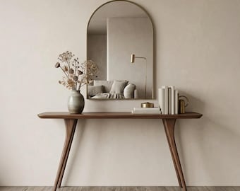 Puede incluir: Una mesa consola de madera oscura con cuatro patas cónicas, sobre la cual hay un jarrón de cerámica con flores secas, una pila de libros blancos y un pequeño objeto metálico. Un espejo de pared arqueado cuelga encima, reflejando una lámpara y un sofá.