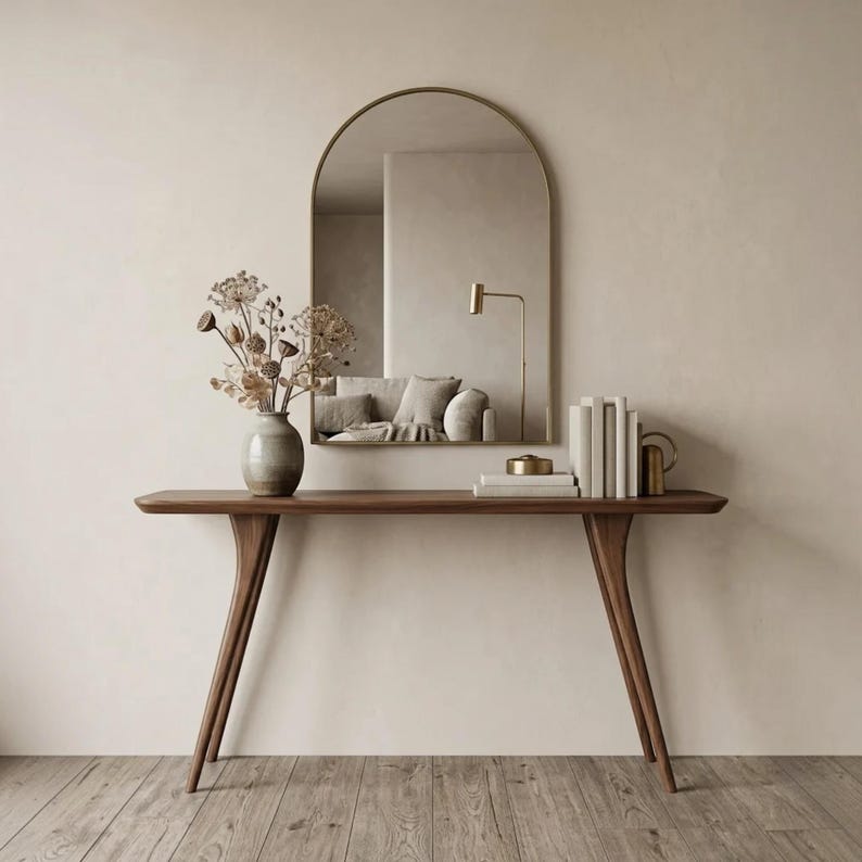 May include: A dark wood console table with four tapered legs, topped with a ceramic vase containing dried flowers, a stack of white books, and a small metallic object. An arched wall mirror hangs above, reflecting a lamp and a sofa.