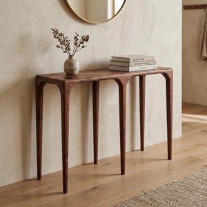 Puede incluir: Una mesa consola de madera marrón oscuro con cuatro patas delgadas y cónicas y un diseño de faldón arqueado. Sobre la superficie hay un pequeño jarrón de cerámica clara con tallos botánicos secos y una pila de tres libros de tapa dura.
