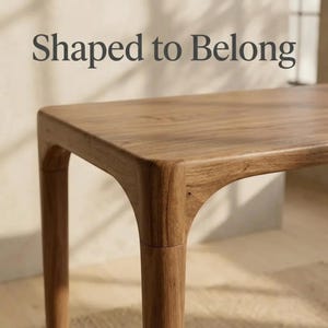 May include: A light brown wooden table with a smooth, rectangular top and rounded corner joints connecting to tapered legs. The text "Shaped to Belong" is printed in dark grey letters above the table surface, which features a visible wood grain.