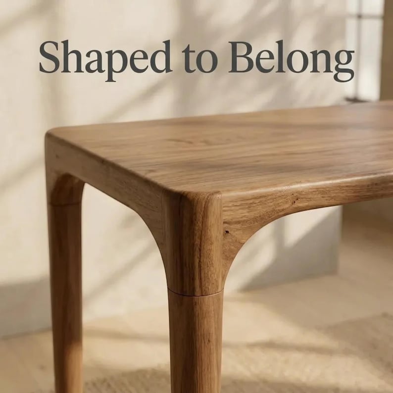 May include: A light brown wooden table with a smooth, rectangular top and rounded corner joints connecting to tapered legs. The text "Shaped to Belong" is printed in dark grey letters above the table surface, which features a visible wood grain.