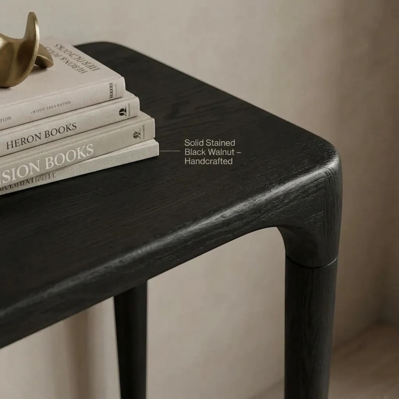 Puede incluir: Una mesa de madera de nogal maciza te&ntilde;ida de negro con bordes redondeados y patas c&oacute;nicas. Una pila de tres libros descansa sobre la superficie, con un objeto dorado sobre el libro superior. El texto en la mesa dice: Solid Stained Black Walnut &ndash; Handcrafted.